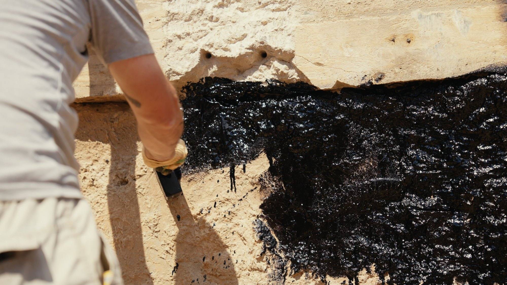 A worker brushing black waterproofing coating onto the outside of a foundation wall